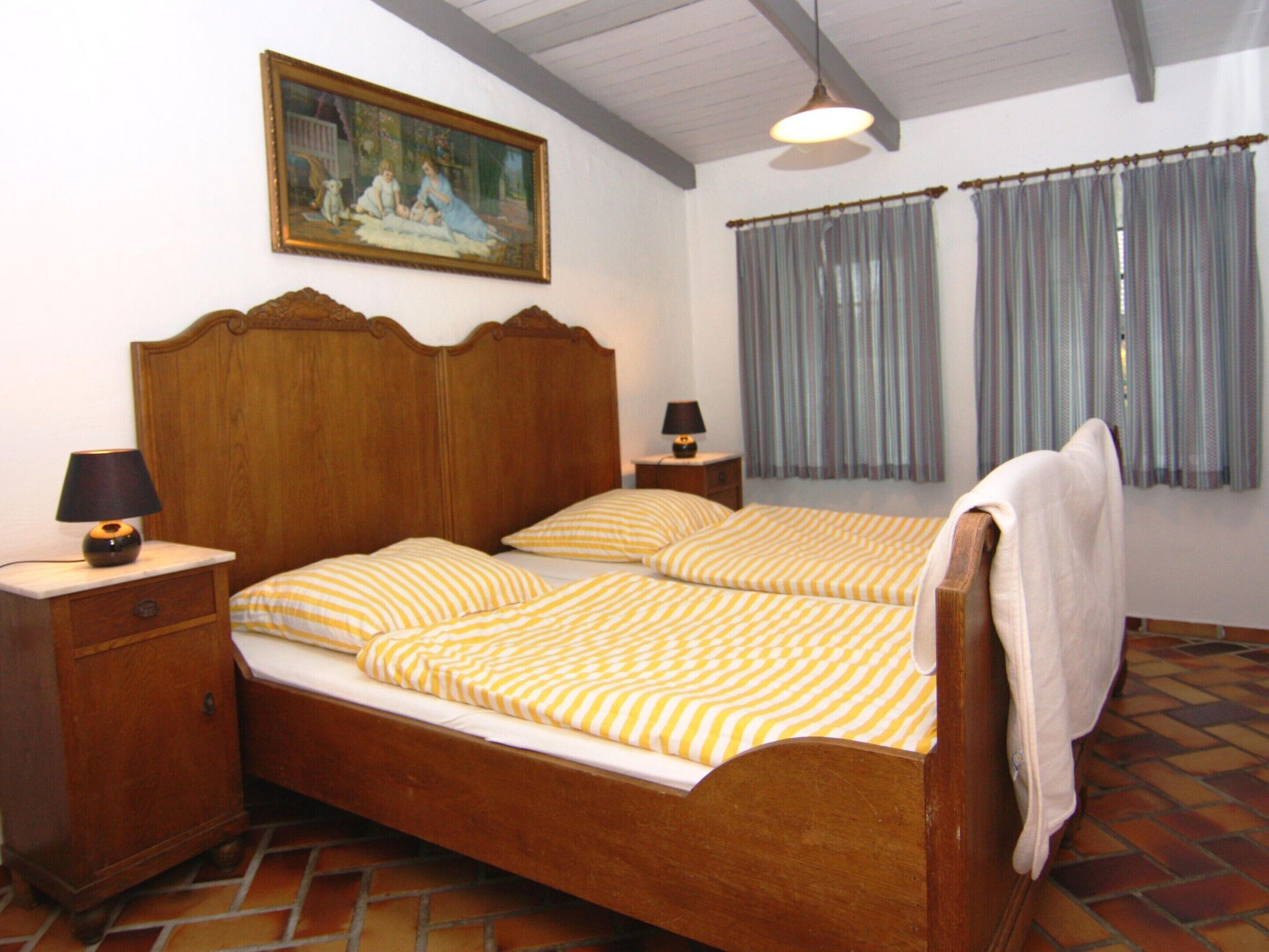 A double bed with a wooden headboard, two bedside tables with lamps, a painting on the wall, and a window with curtains.