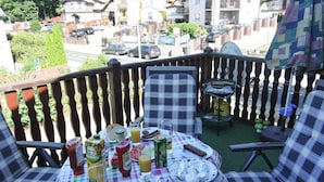 Un balcon avec une nappe à carreaux, des sièges d’extérieur et une vue sur une rue bordée de voitures garées et d’immeubles.