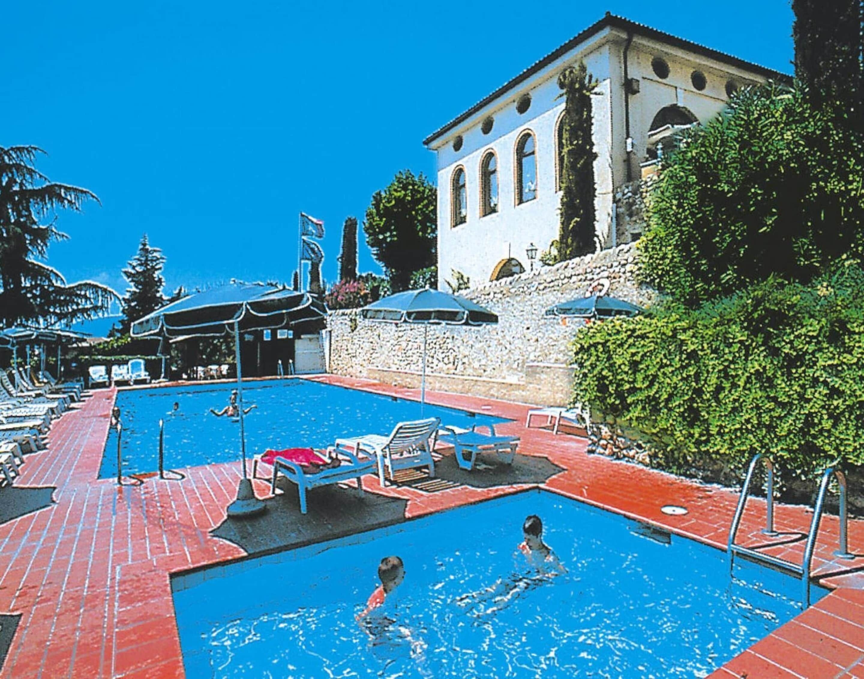 Ein Poolbereich mit Liegestühlen und Sonnenschirmen, eine Steinmauer sowie ein Gebäude mit Bogenfenstern.