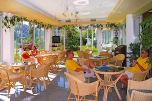 A sunlit indoor seating area with wicker furniture and large windows offering views of greenery.