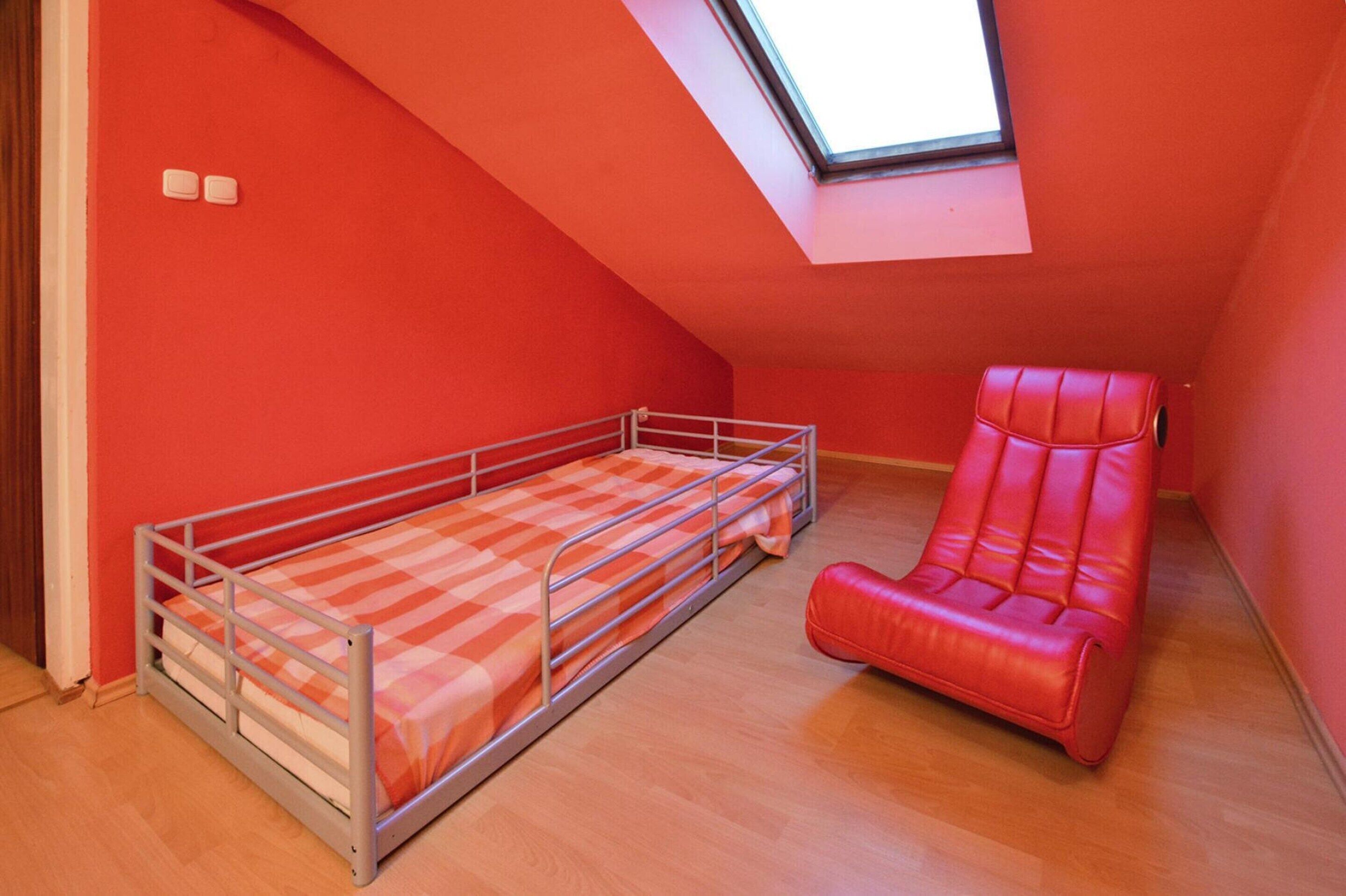 A small, red-walled room with a single metal bed and a red chair.