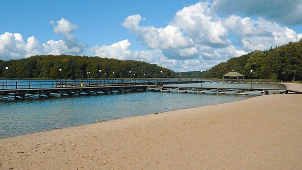 A beach with a pier extending into calm water, surrounded by trees and a clear sky. - Comfy Lakeside House With Private Garden (Ińsko)