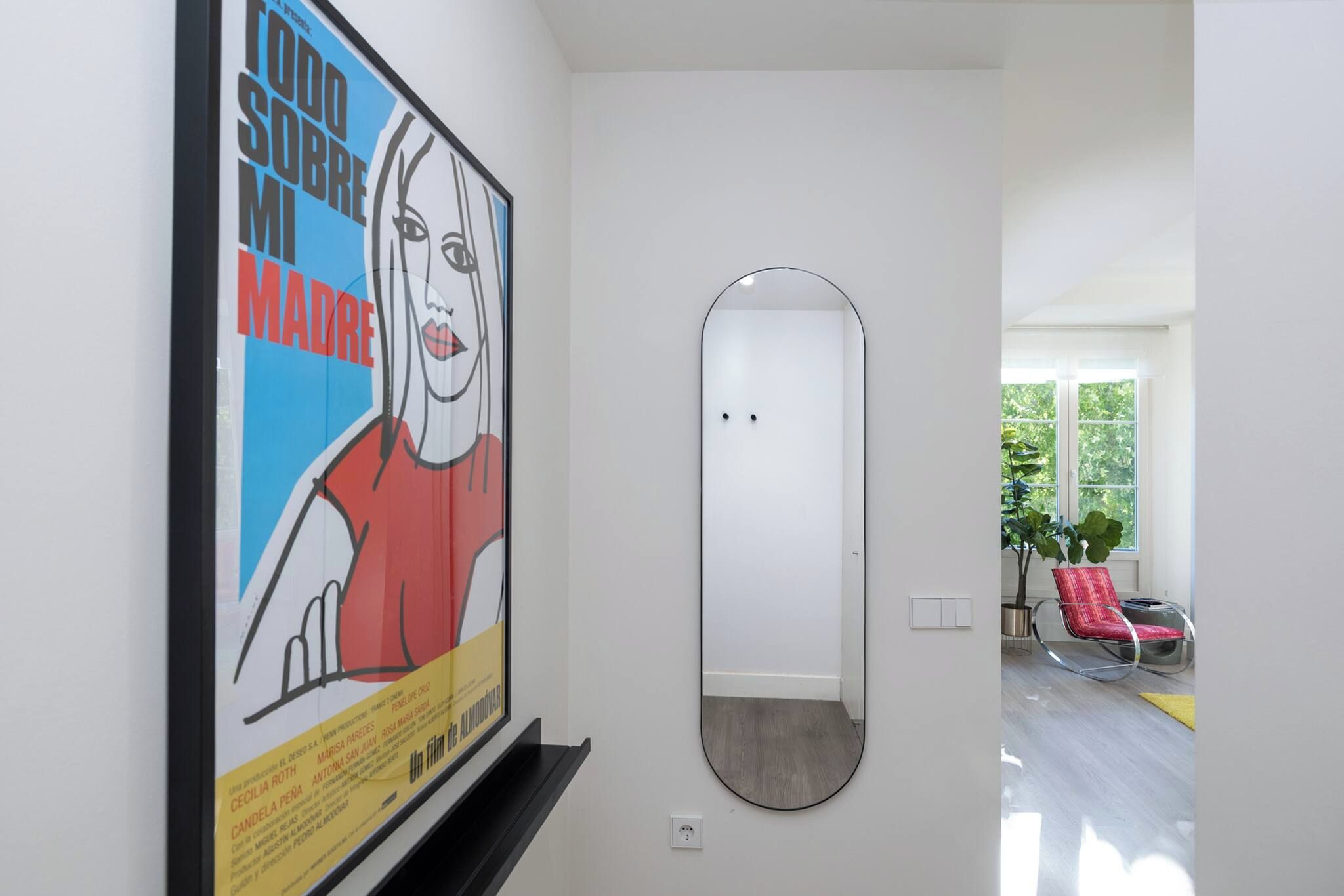 A hallway with a poster, a mirror, and a view into a room with a chair and a plant.