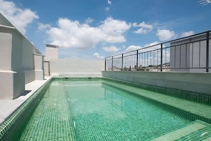 Una piscina en la azotea con agua azul cristalina, rodeada por un edificio blanco con barandilla de vidrio.