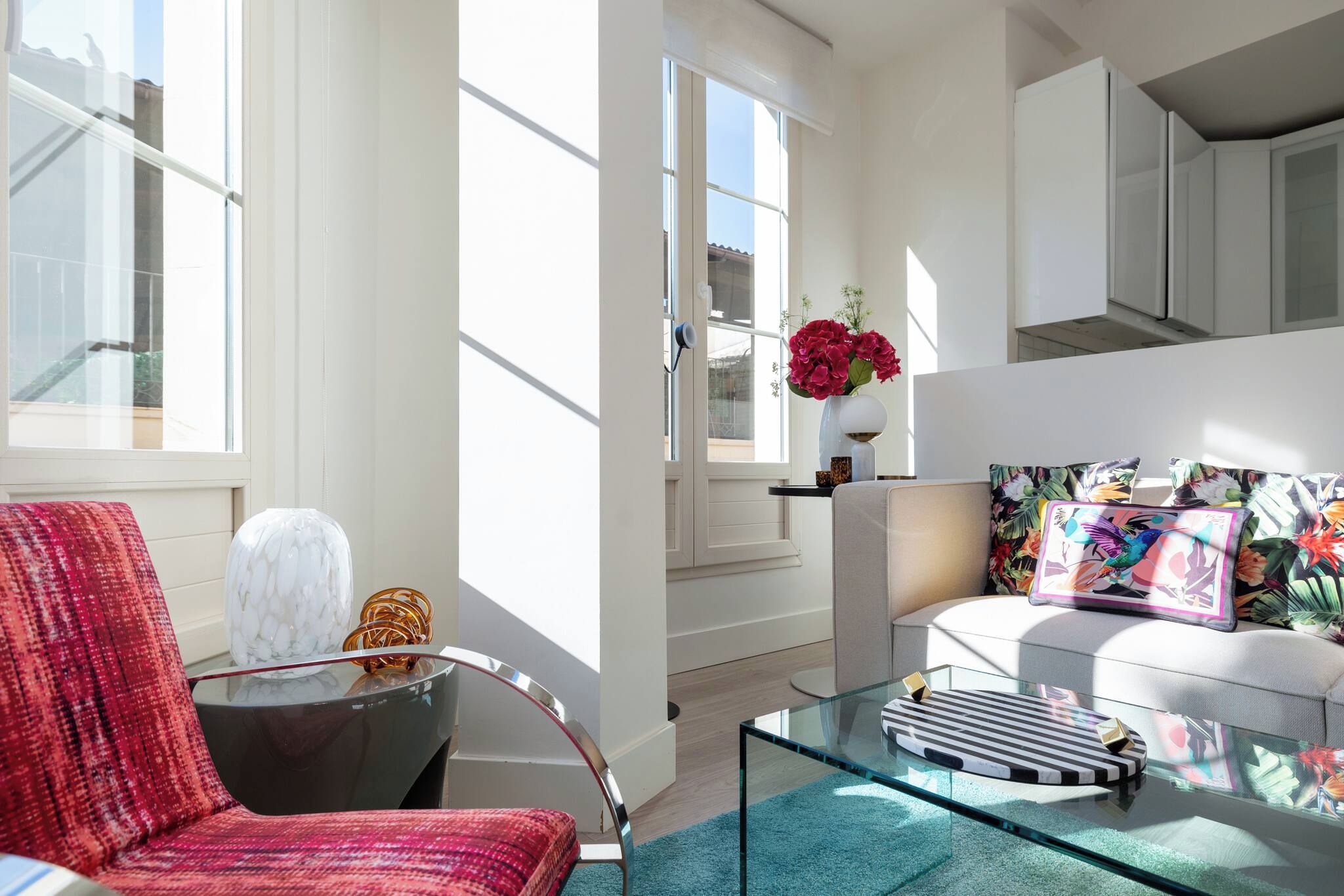 Een moderne woonkamer met een glazen salontafel, een rode fauteuil en een bank met kleurrijke kussens.