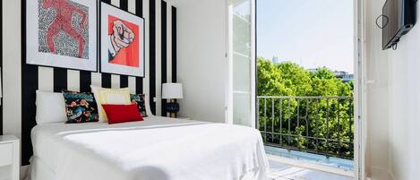 A bedroom with a large bed, two bedside tables, a window, and a balcony with a view of trees and buildings.