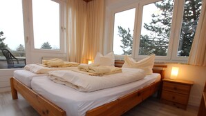 A wooden bed with white linens and pillows, a bedside table with a lamp, and large windows with curtains.