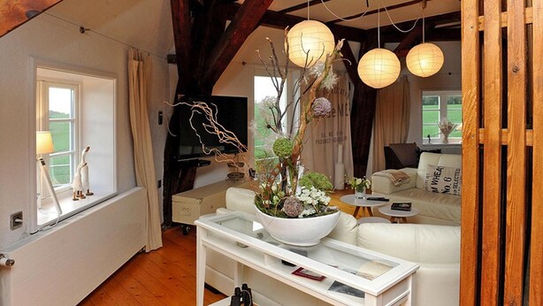 A cozy living room with exposed wooden beams, a white sofa, a coffee table with a floral centerpiece, and hanging paper lanterns. - Hollaender Windmuehle in Neubukow (Neubukow)