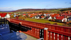 En balkong med ett rött räcke som vetter ut mot en by med hus som har röda tak och ett grönskande landskap.