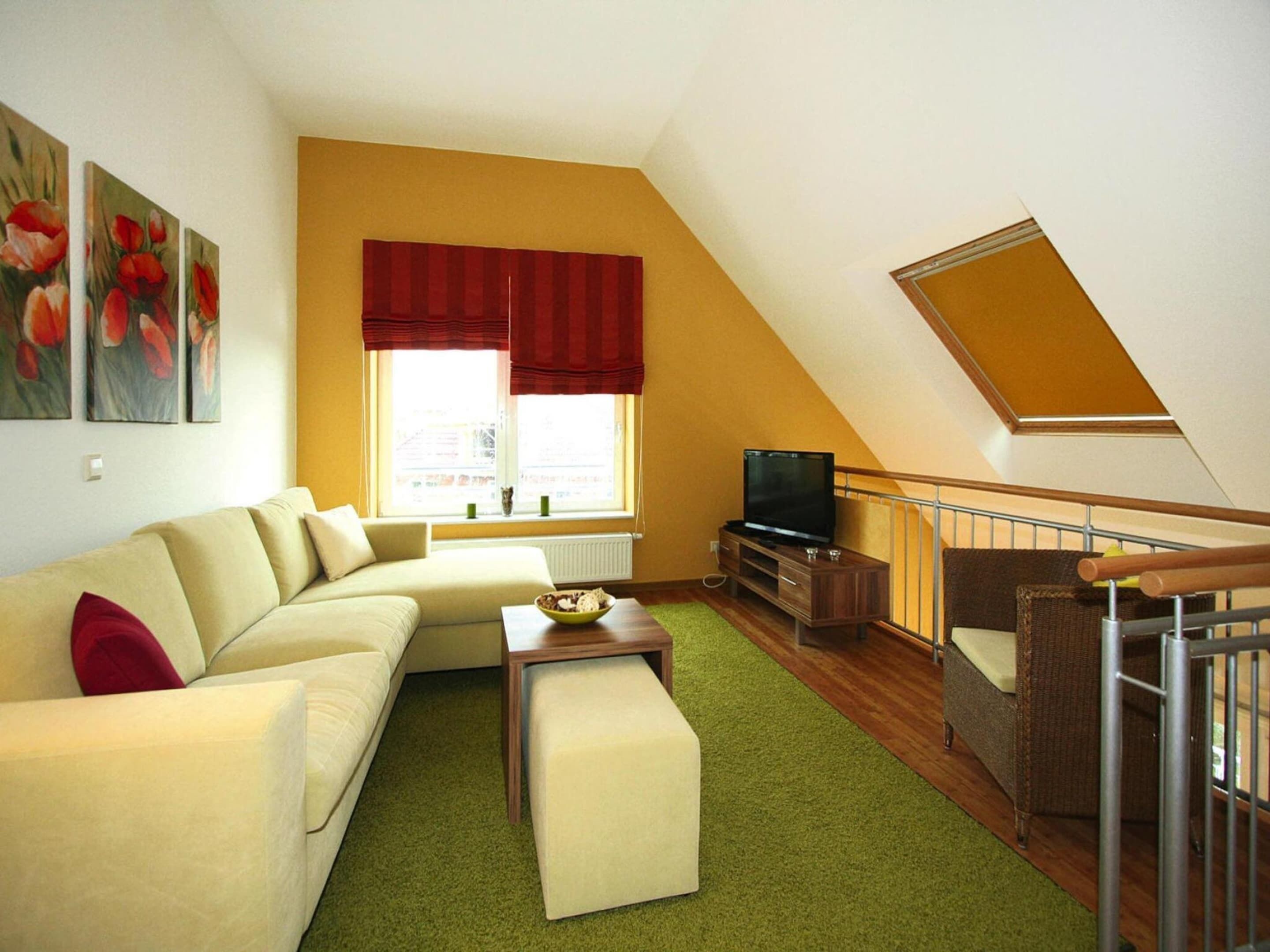 A living room with a sofa, ottoman, and a television on a wooden stand. There is a window with red curtains and a painting of flowers on the wall.