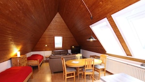 A cozy attic bedroom with a loft bed, a sofa, a dining table with chairs, a TV, and a large window providing natural light. - Ferienwohnung in Westerland auf Sylt (Sylt)