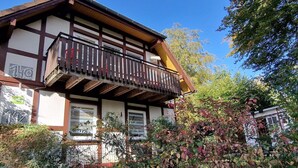 A traditional chalet with a wooden balcony, surrounded by dense foliage and a clear blue sky. - Ferienwohnung in Frankenau (Frankenau)