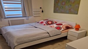 A bedroom with a bed, a bedside table, a lamp, a radiator, a window with blinds, and a wall painting. - Ferienwohnung in Frankenau (Frankenau)