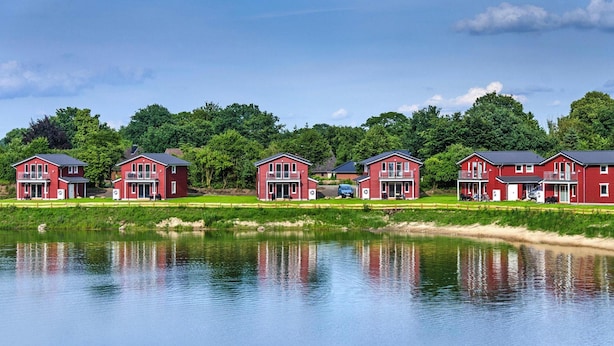 A row of red buildings with balconies, a pond in the foreground, and trees in the background. - Ferienhaus in Seenhhe in Hechthausen (Hechthausen)