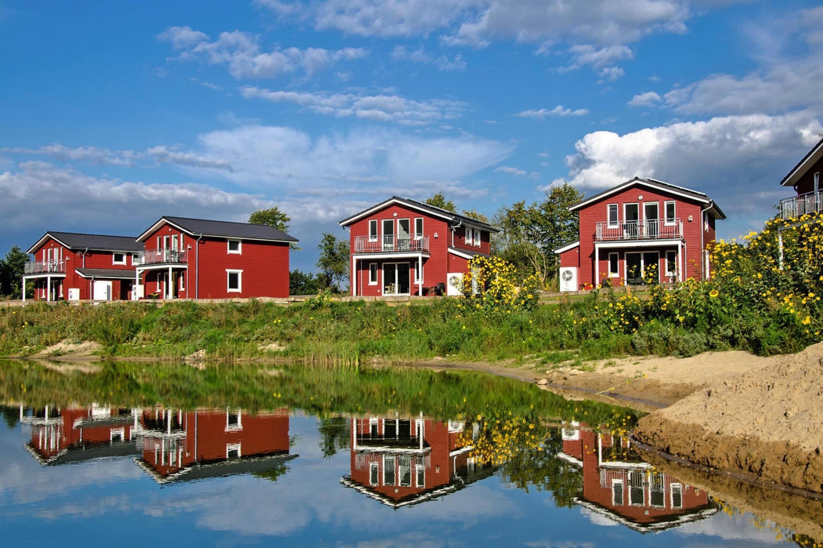 Une rangée de maisons rouges dotées de balcons, qui se reflètent dans une étendue d’eau calme, le tout entouré de verdure et de fleurs jaunes.