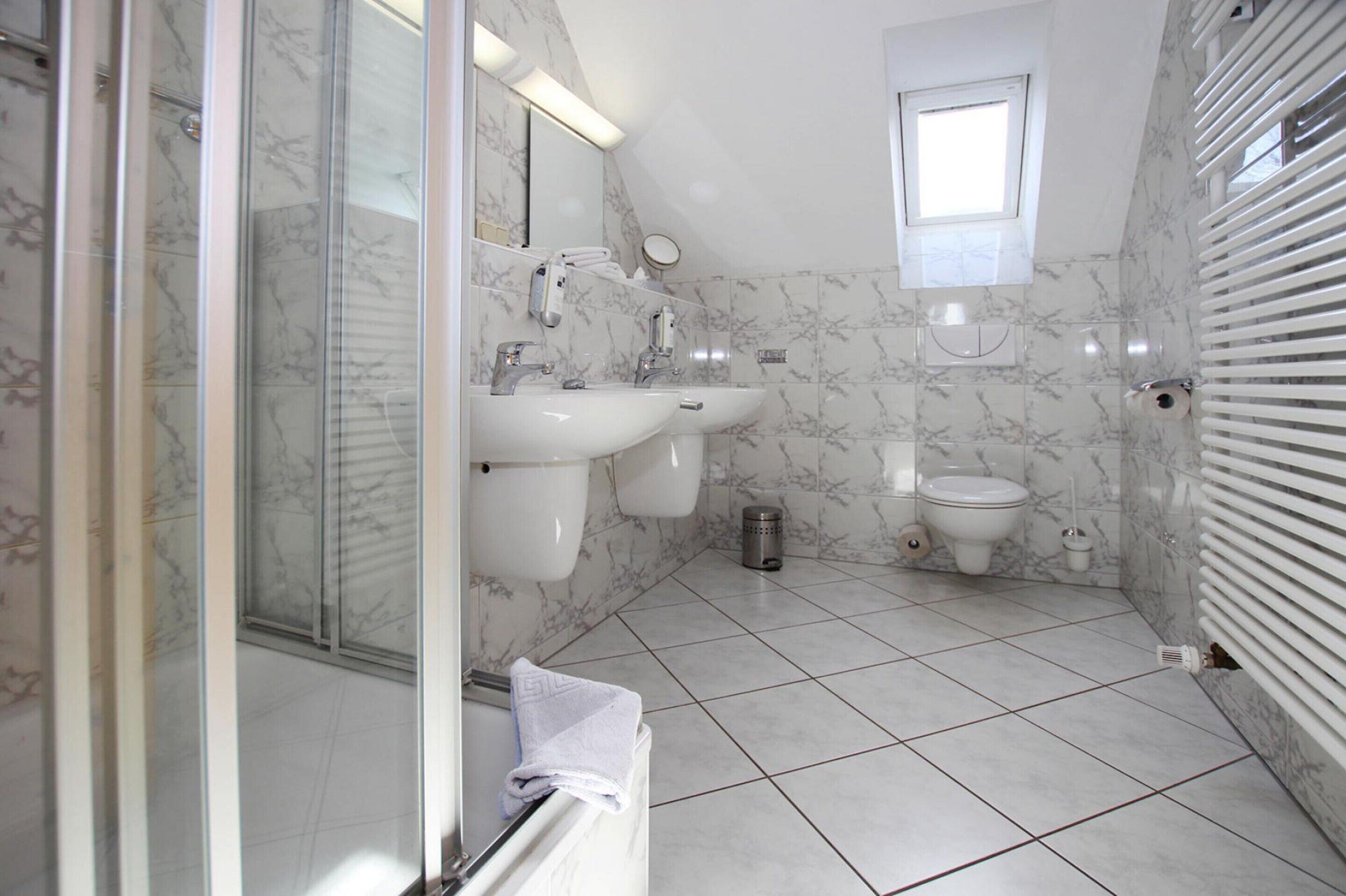 A bathroom with a glass shower enclosure, two sinks, a toilet, and a small window.