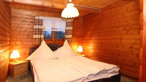 Une chambre dans une cabane en bois, avec un lit, deux lampes de chevet, une fenêtre avec des rideaux à motifs et un luminaire suspendu.