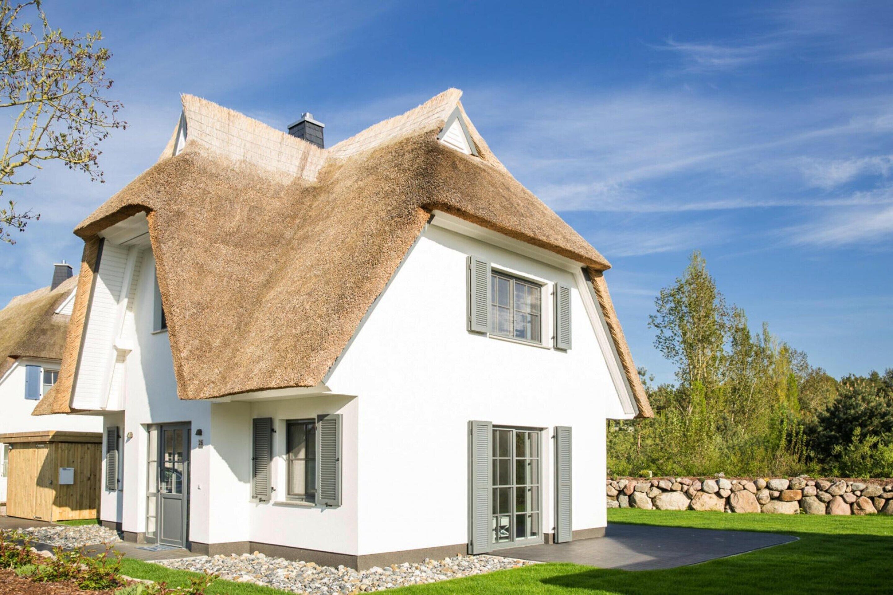 A white house with a thatched roof and shutters, surrounded by a stone wall and greenery.