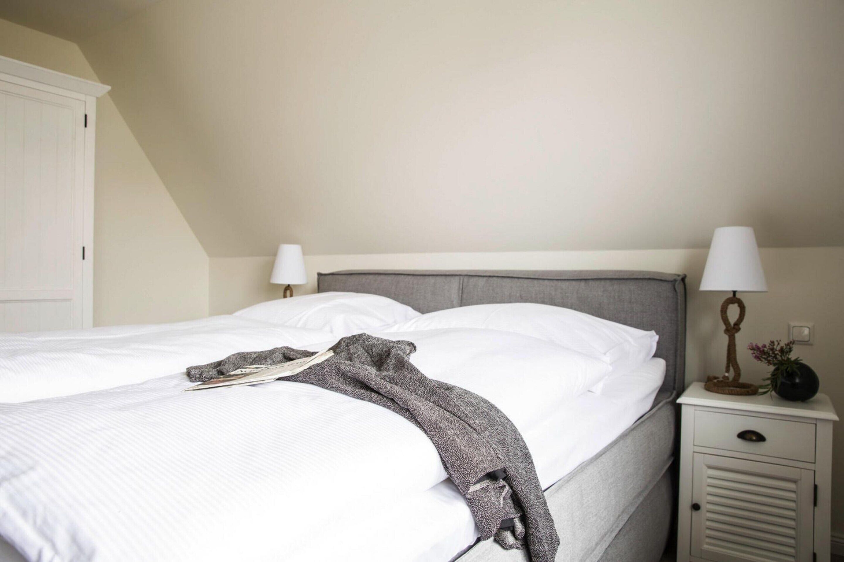 A neatly made bed with white linens, a grey throw blanket, and a bedside table with a lamp and a small decorative item.