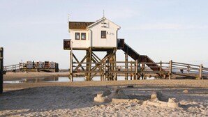 A beach house with a wooden deck, a small staircase, and a sandy beach in the foreground. - Ferienwohnung, St. Peter-ording (St. Peter-Ording)