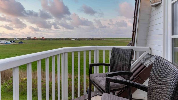 A balcony with wicker furniture and a white railing overlooking a grassy field and a cloudy sky. - Ferienwohnung, St. Peter-ording (St. Peter-Ording)