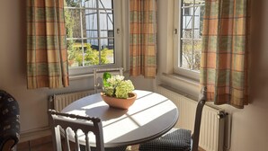 A corner room with two windows, a radiator, a round table with a bowl of flowers, and a chair with a patterned cushion. - Gemutliche Ferienwohnung in Fassberg (Faßberg)