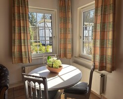 Habitación de esquina con dos ventanas, un radiador, una mesa redonda con un cuenco de flores y una silla con un cojín estampado.