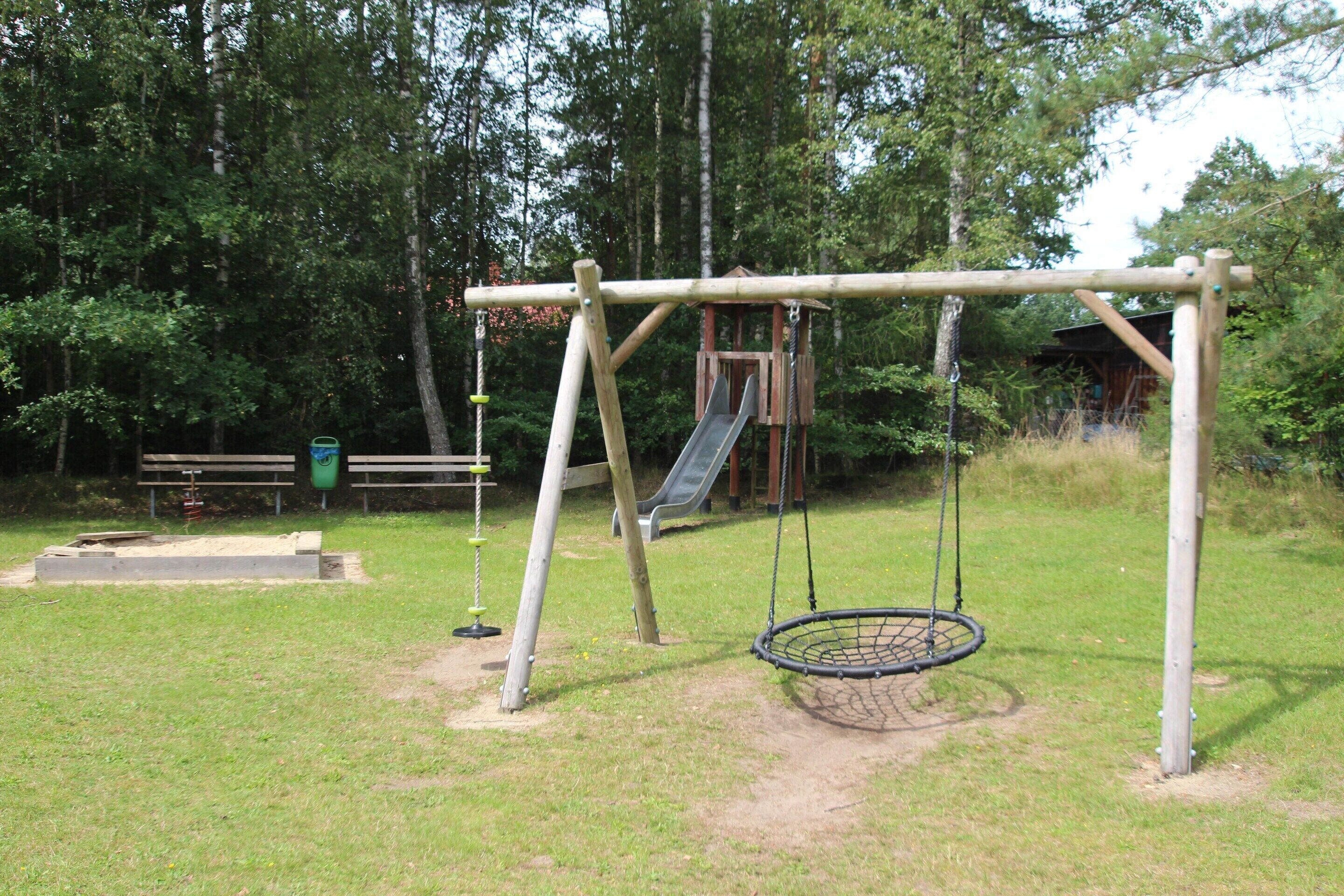 Un parque infantil con columpios y tobogán, rodeado de árboles.