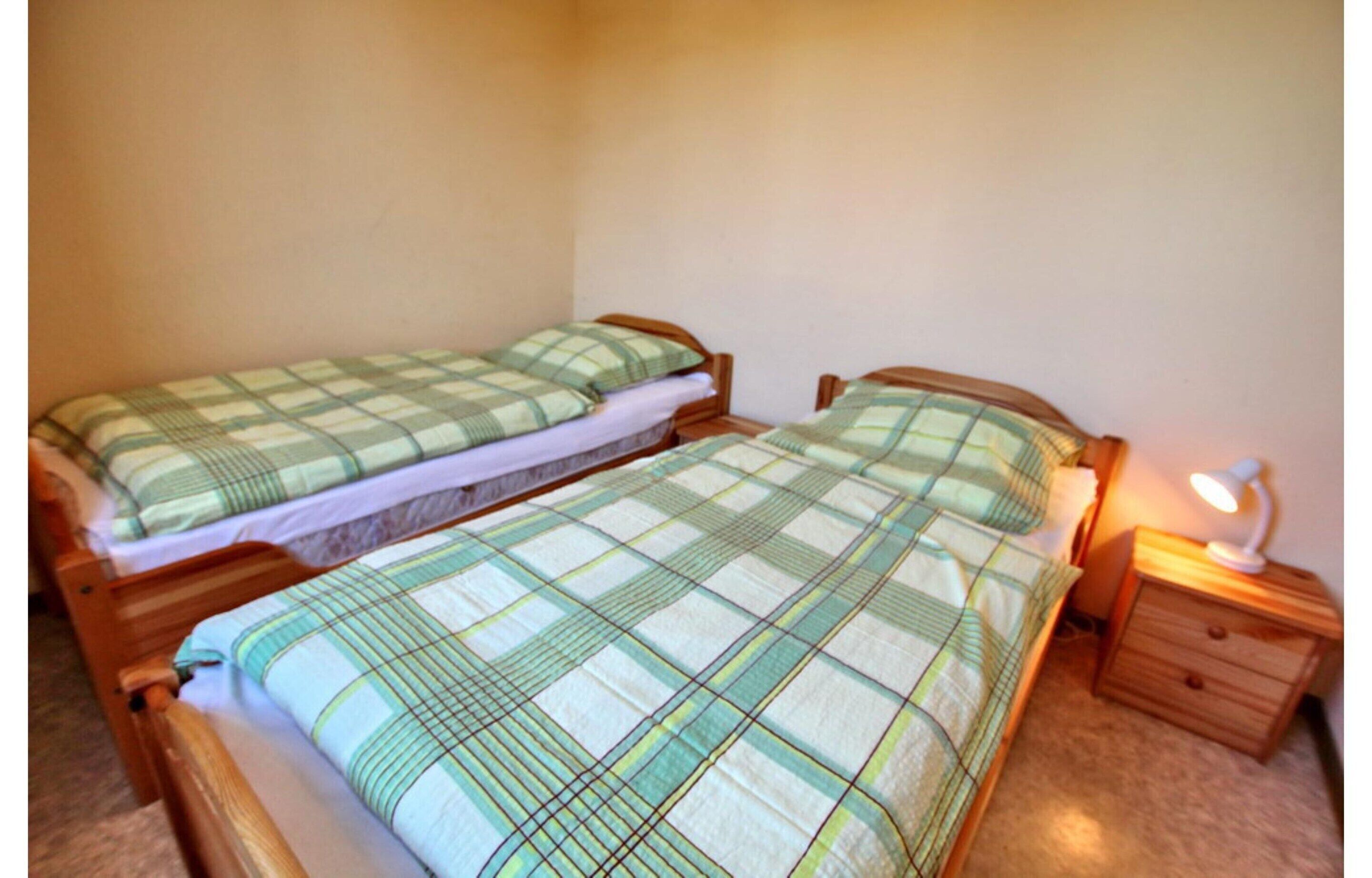 Um quarto pequeno e acolhedor com duas camas de solteiro, cada uma com estrutura de madeira e colcha xadrez. Há uma mesa de cabeceira com uma luminária de um lado.