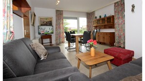A living room with a grey sofa, a wooden coffee table with a vase of flowers, a dining area with a table and chairs, and a kitchen area with wooden cabinets. - Bungalow Near Dornumersiel Beach (Dornum)