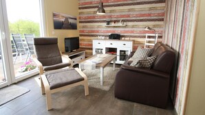 Un salon comprenant un canapé, un fauteuil, un téléviseur et une table basse en bois. Une fenêtre offre une vue sur la verdure à l’extérieur.