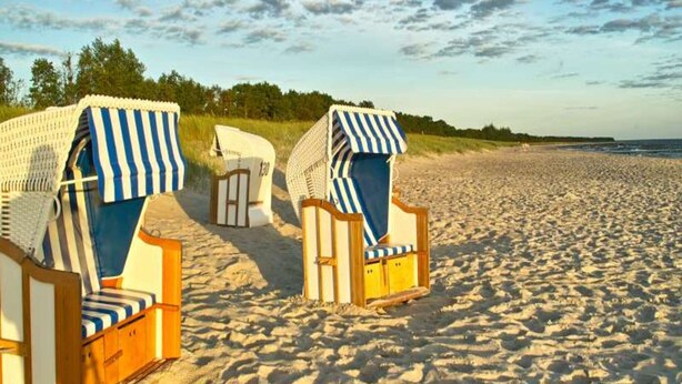 Two beach chairs with striped covers on a sandy beach, a wooden bench, and a gate labeled "130" in the background. - Ferienhaus, Fuhlendorf (Fuhlendorf)