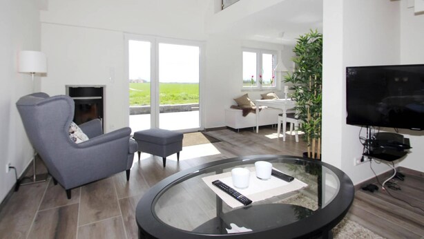 Ett modernt vardagsrum med ett soffbord i glas, en grå soffa, en platt-TV och ett stort fönster med utsikt över ett grönskande landskap.