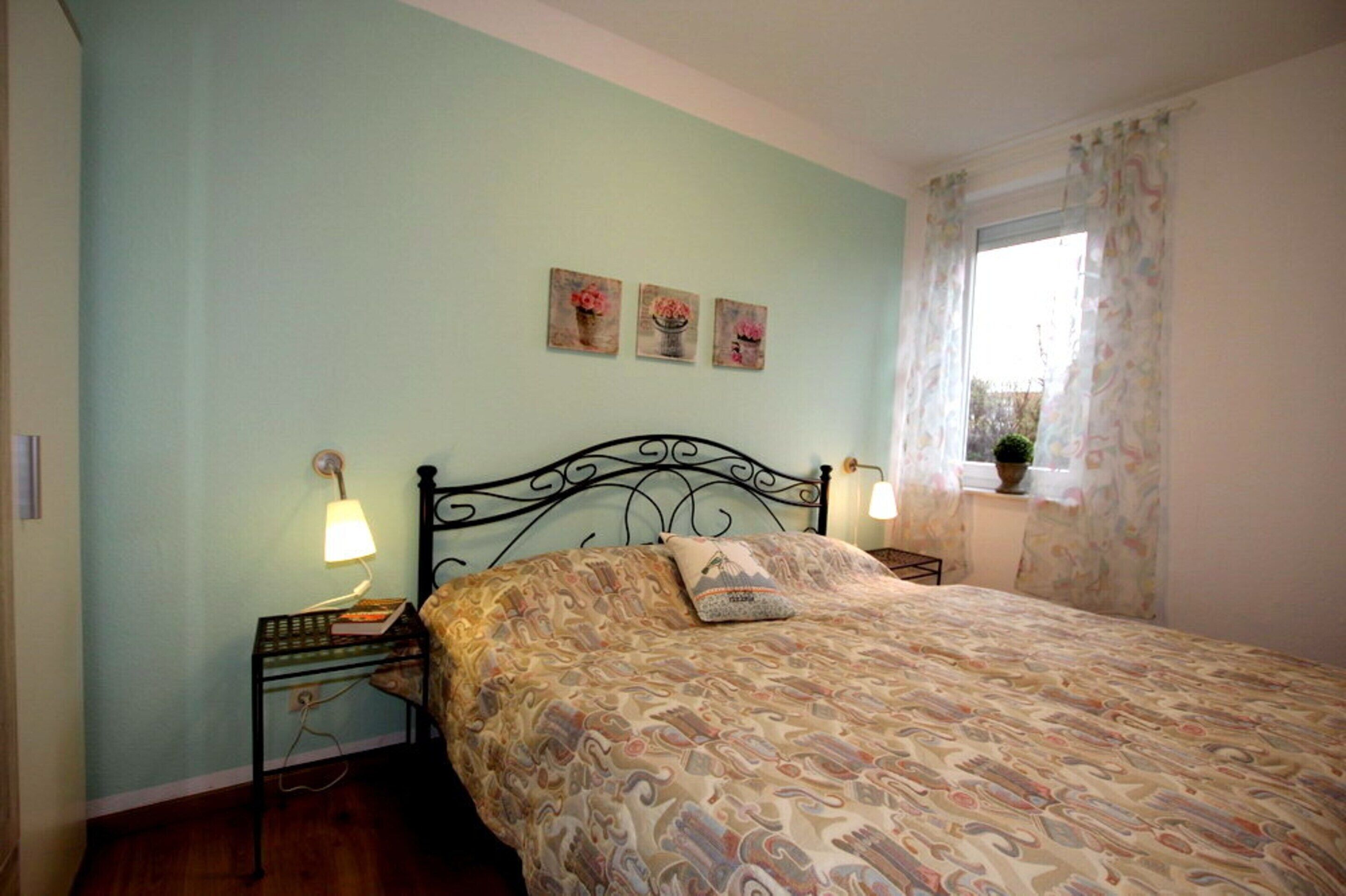 A bedroom with a bed, two bedside lamps, a nightstand, and three floral paintings on the wall.