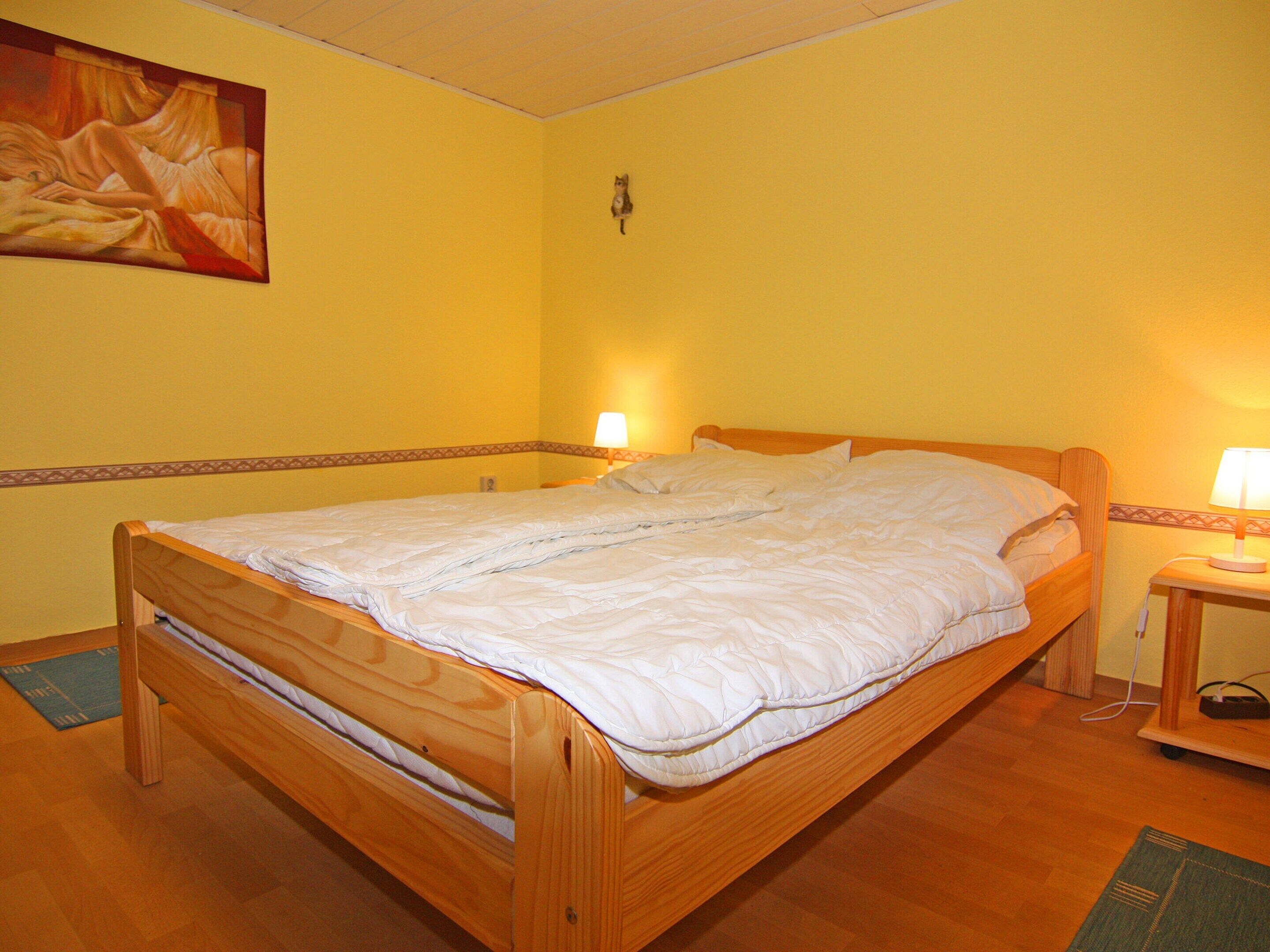 Une chambre avec un lit simple, un cadre en bois, une couvre-lit blanche, une table de chevet avec une lampe et un tableau au mur.