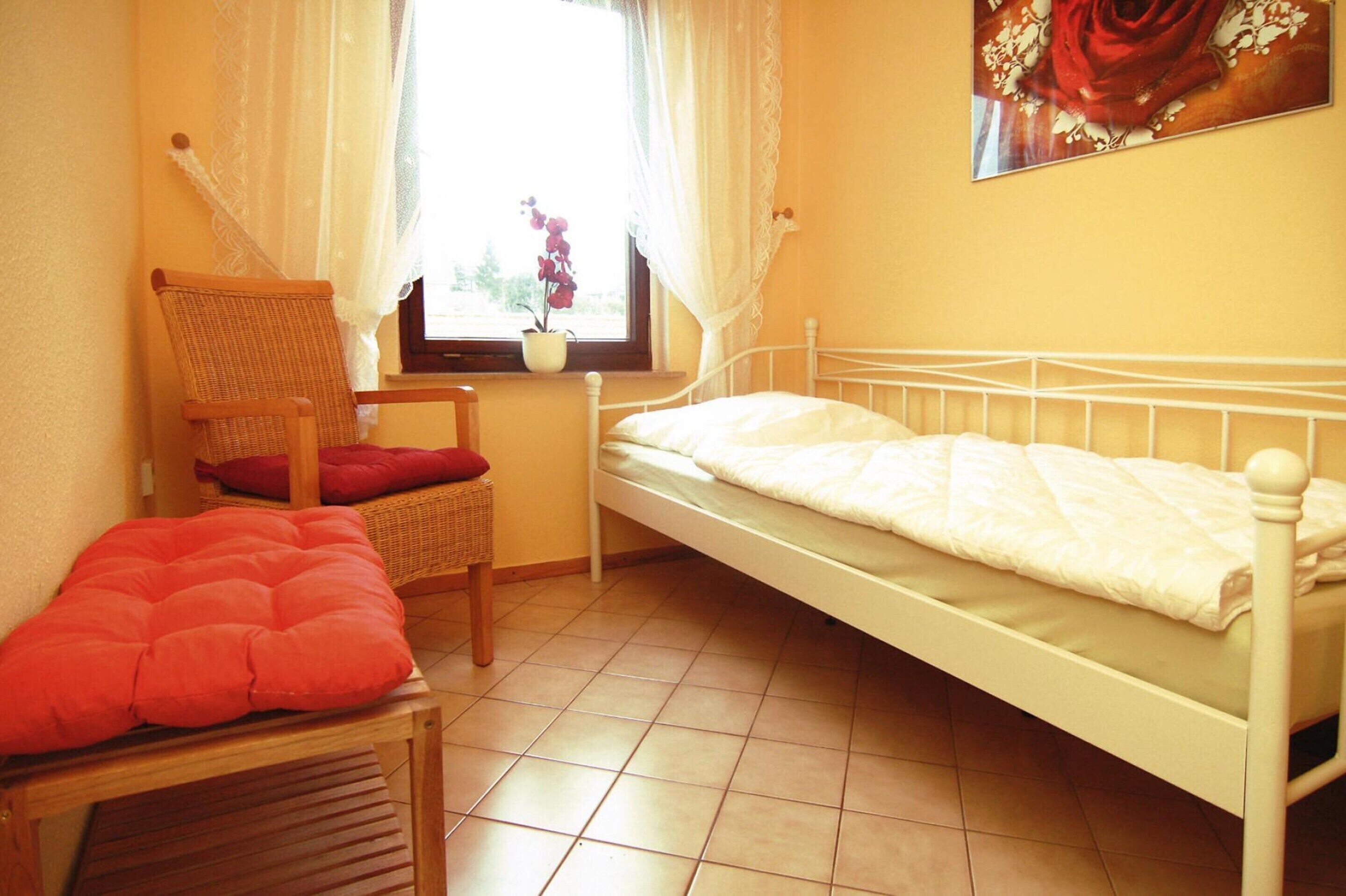 Een slaapkamer met een enkel bed, een stoel met een rode kussen, een kleine tafel en een raam met gordijnen.