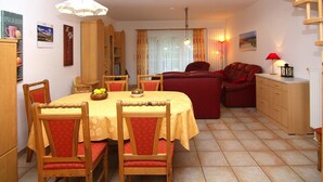 Un comedor con una mesa dispuesta para cuatro personas, rodeada de sillas de madera con cojines rojos. En el fondo, un sofá rojo y un aparador de madera.