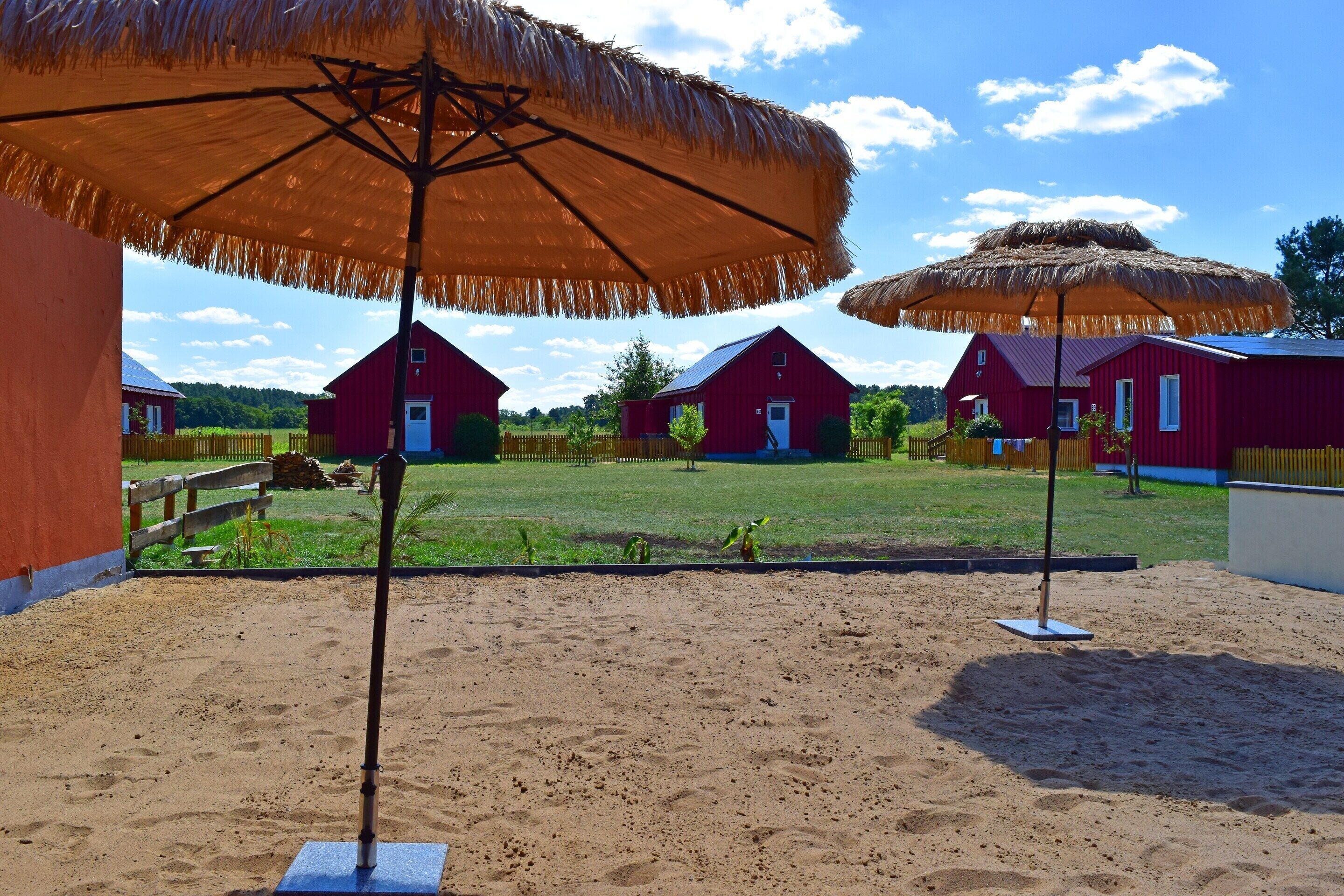 A sandy outdoor area with two thatched umbrellas and red barns in the background.