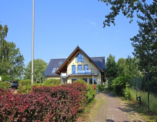Une maison à deux étages, dotée d’un toit en pente, d’un balcon et d’une allée pavée menant à l’entrée.