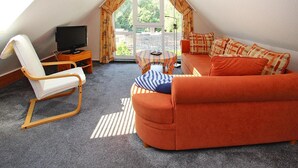 Um quarto de sótão acolhedor, com teto inclinado, uma cadeira branca, um sofá, uma televisão e vista para o verde lá fora.