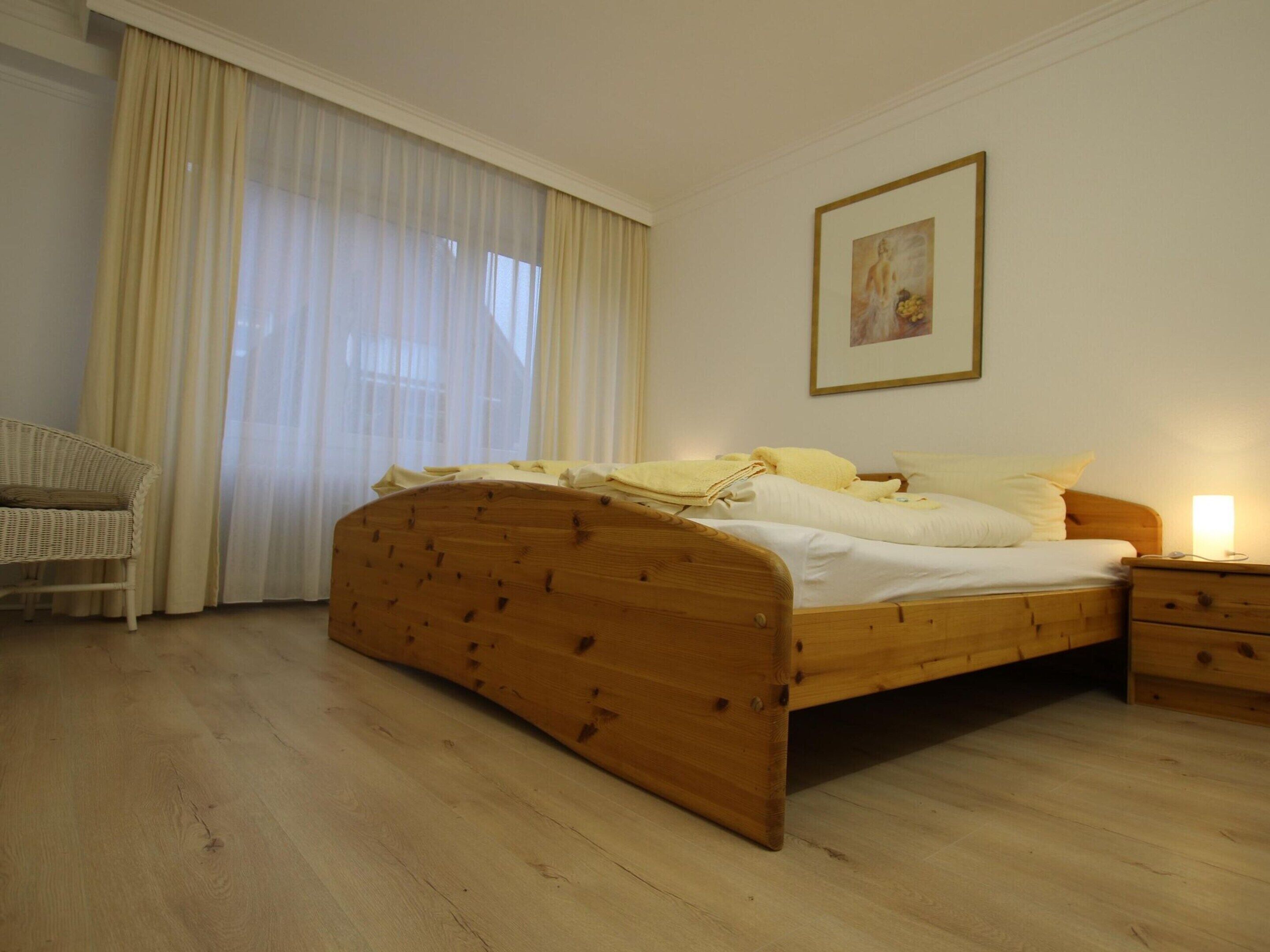 Een slaapkamer met een houten bed, een nachtkastje, een rieten stoel en een groot raam met dunne gordijnen.