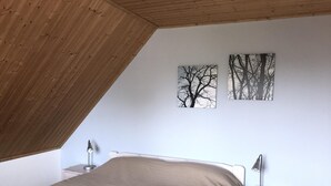 A bedroom with a sloped ceiling, a bed with a beige bedspread, two bedside tables with lamps, and two framed pictures on the wall. - Ferienhaus Christina, Burhave (Butjadingen)