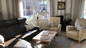 A living room with a black leather sofa, two beige armchairs, a wooden coffee table with woven placemats, a floor lamp, and a window with curtains. - Ferienhaus Christina, Burhave (Butjadingen)