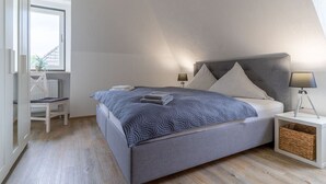 Une chambre moderne avec un plafond en pente, un grand lit doté d’une tête de lit grise, deux tables de chevet avec des lampes, un fauteuil et une vue sur l’extérieur par une fenêtre.