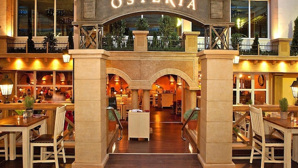 A hotel lobby with an arched entrance labeled "OSTERIA," featuring a central staircase, seating areas, and a view of the outdoor area. - Appartements im Freizeitpark Weissenhauser Strand (Wangels)
