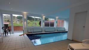 En indendørs swimmingpool med fliset gulv, en bænk langs væggen og udsigt til en have gennem store vinduer.