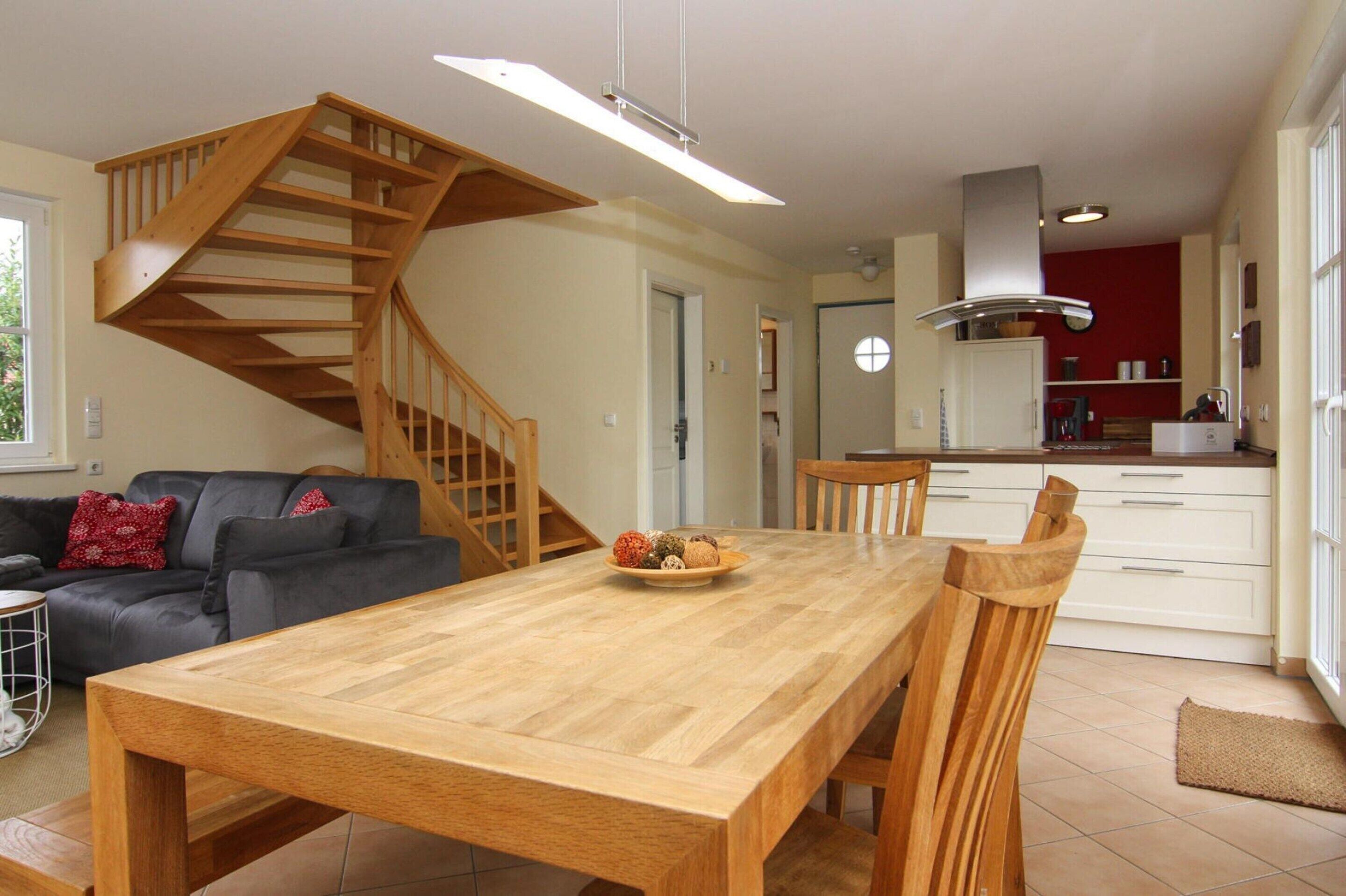 A spacious living area with a wooden staircase, a dining table with chairs, a sofa, and a kitchen area with white cabinets and a red accent wall.