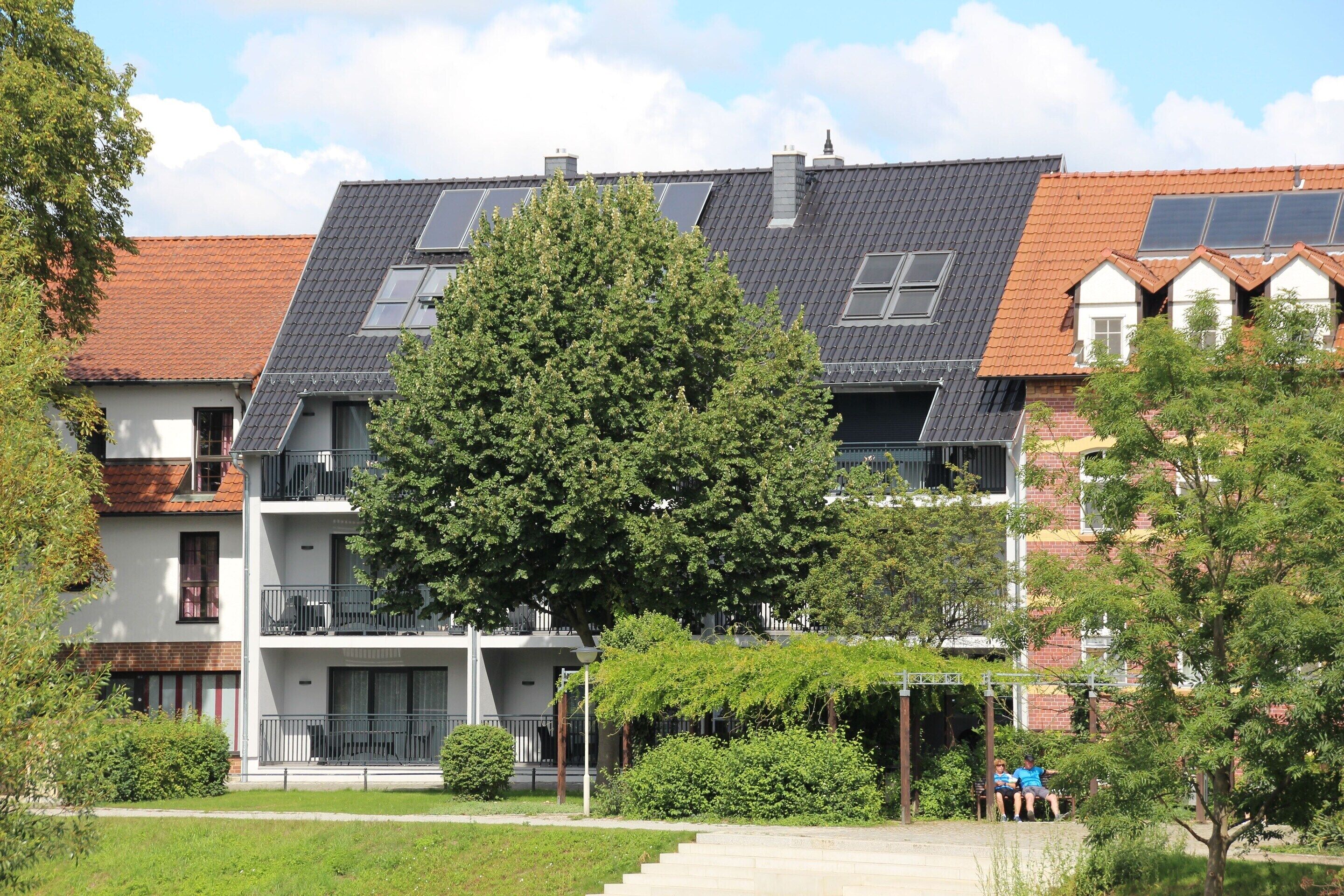 A multi-story residential building with balconies, solar panels, and a large tree in the foreground.
