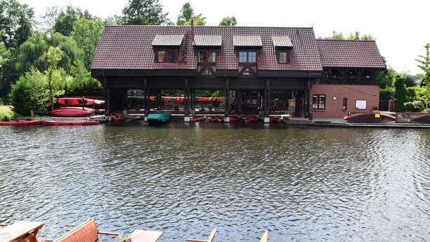 A riverside building with a covered deck area, boats docked nearby, and wooden benches along the water's edge. - Appartements mit Blick auf die Spree in Lubben (Lübben (Spreewald))