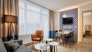 A modern hotel room with a grey sofa, a dining table with chairs, a TV mounted on a patterned wall, and a large window with sheer curtains. - Appartements im Freizeitpark Weissenhauser Strand (Wangels)